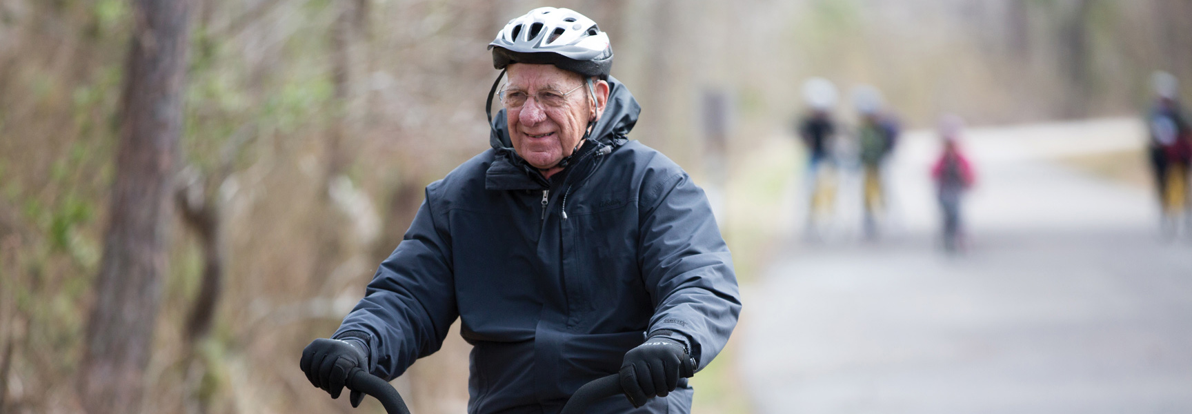An older man in a helmet smiles while riding his bicycle on a paved trail through a wooded park in Virginia.