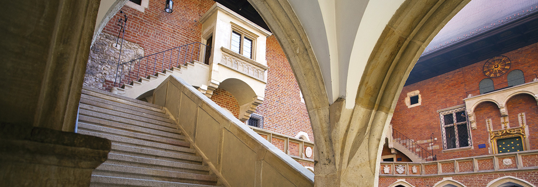 A view through stone arches into a historic brick courtyard with a grand staircase in Poland.