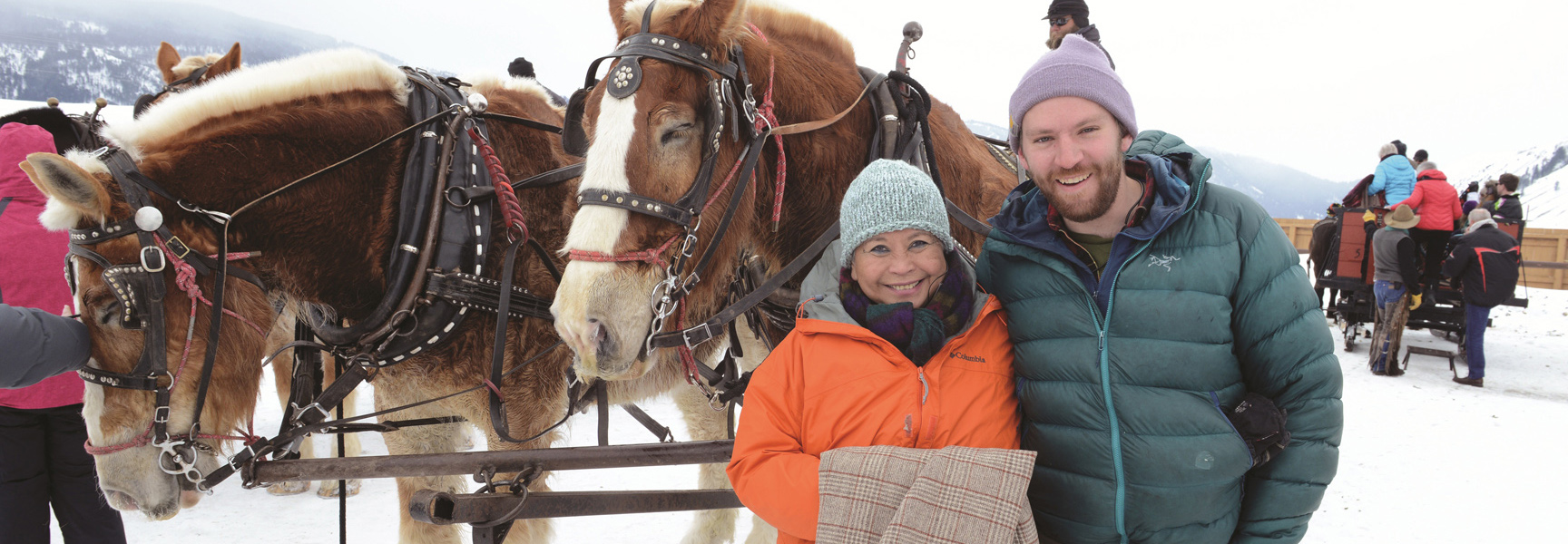 A man and woman smile in front of two harnessed draft horses during a winter sleigh ride in Wyoming.