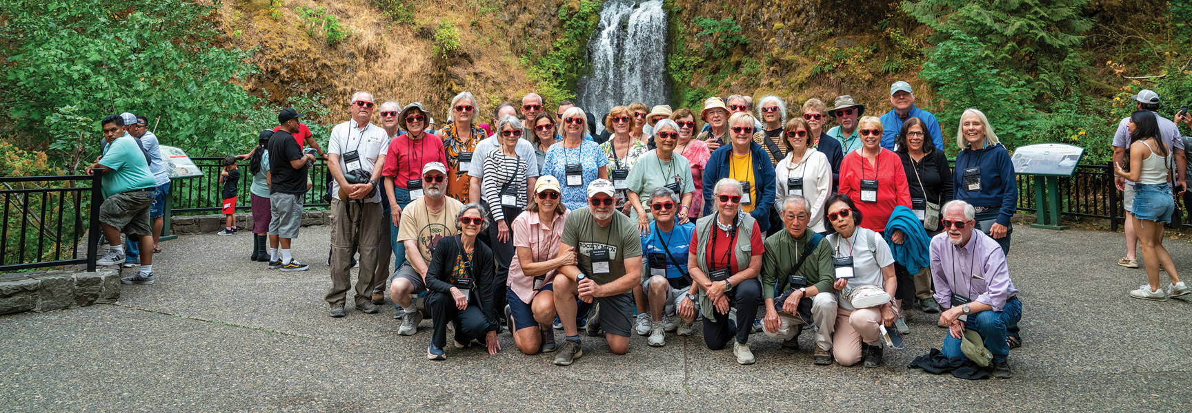 A large tour group wearing matching red sunglasses poses for a photo in front of a scenic waterfall in Oregon.