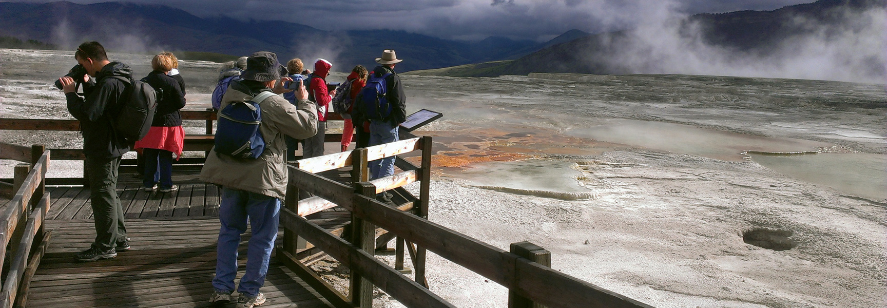 A group of visitors on a wooden boardwalk look out over the vast, steaming geothermal landscape of Yellowstone National Park.