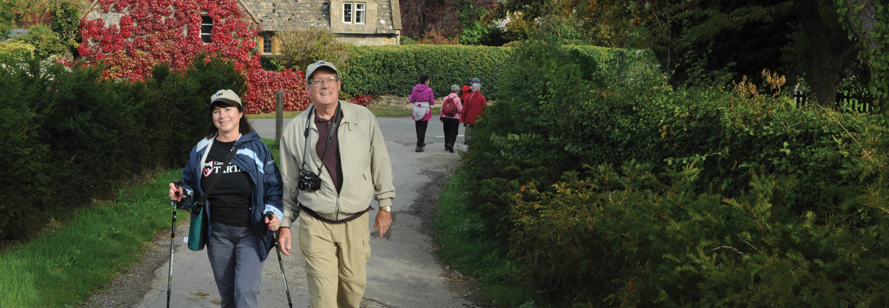 An older couple smiles at the camera while hiking on a path through the countryside in England.