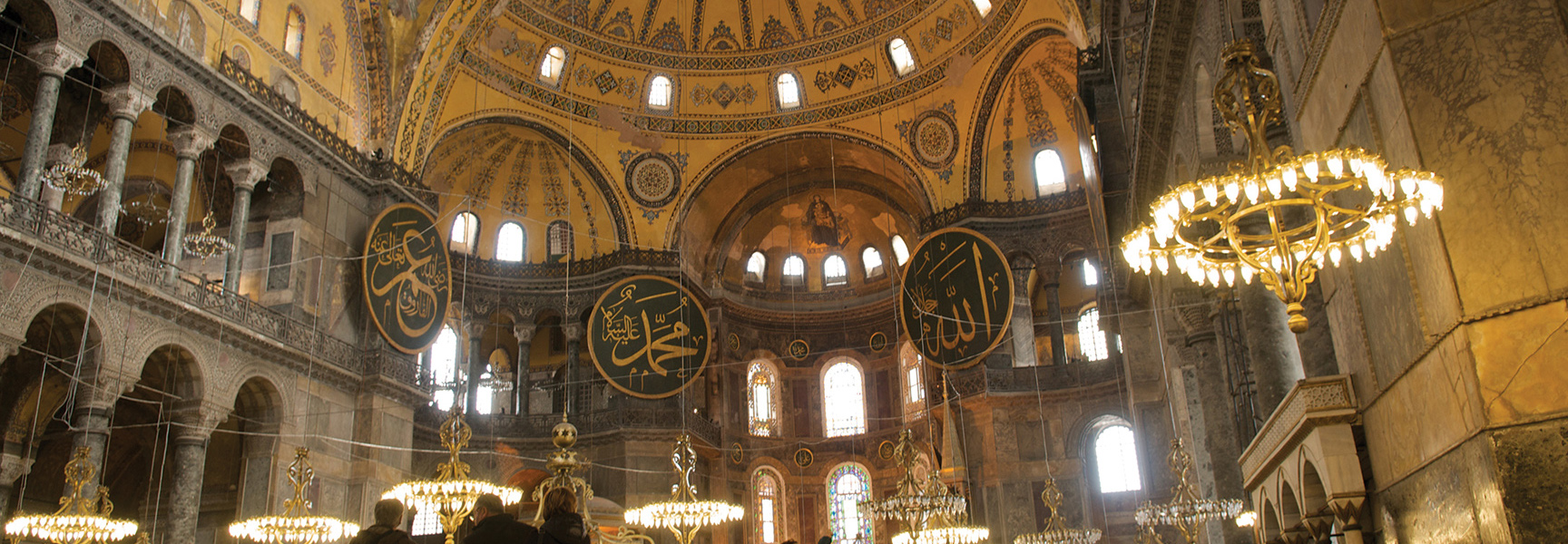 The grand interior of a historic mosque in Turkey, with its massive decorated dome, ornate chandeliers, and large Arabic calligraphy medallions.