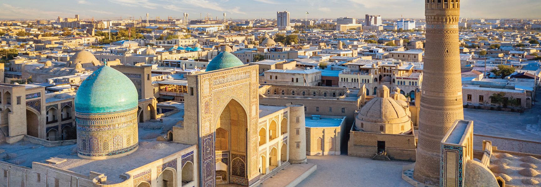 An aerial view of a historic Silk Road city in Central Asia, with turquoise domes and an ornate minaret under a warm sky.