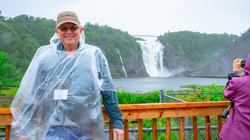 A smiling man in a rain poncho stands on a wooden deck overlooking the large Montmorency Falls Park in Québec.
