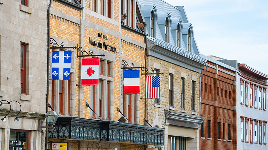 The historic stone facade of Hôtel Manoir Victoria in Old Québec City features several international flags hanging above its entrance.