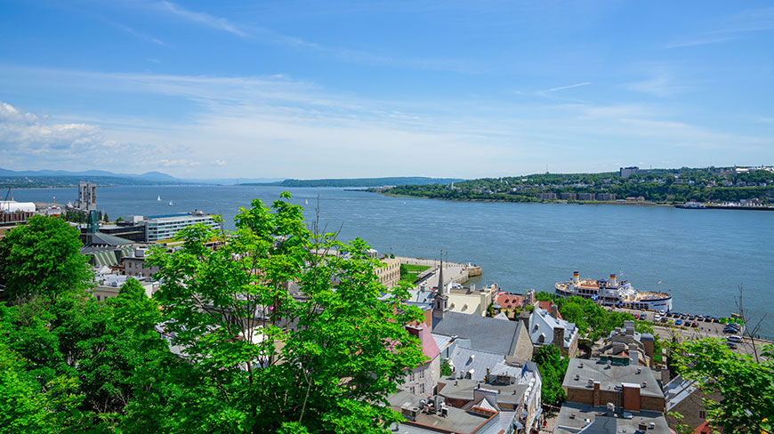 A scenic view of the historic architecture and lush greenery of Old Québec overlooking the St. Lawrence River under a clear blue sky.