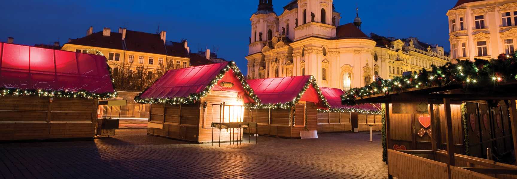 A festive New Year's Eve market in Prague, Czech Republic, with red-roofed stalls and historic buildings lit up at night.