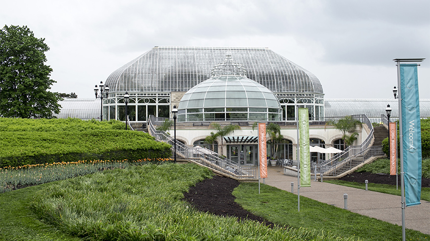 The large glass conservatory at Phipps in Pittsburgh, Pennsylvania, with manicured green lawns and gardens in the foreground under an overcast sky.
