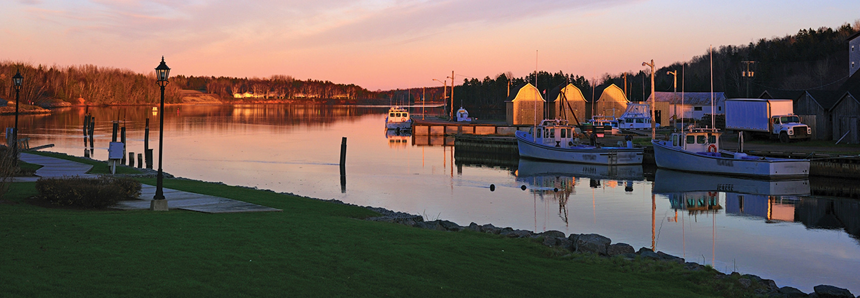 Fishing boats are docked in a tranquil harbor in the Canadian Maritimes at sunset, the calm water reflecting the warm colors of the sky.