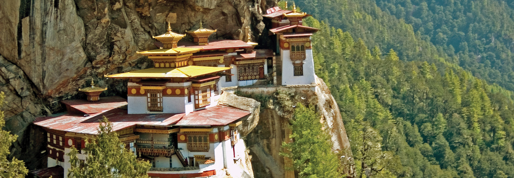 A traditional Bhutanese monastery with white walls and tiered roofs clings to a cliffside, overlooking a lush, forested mountain.