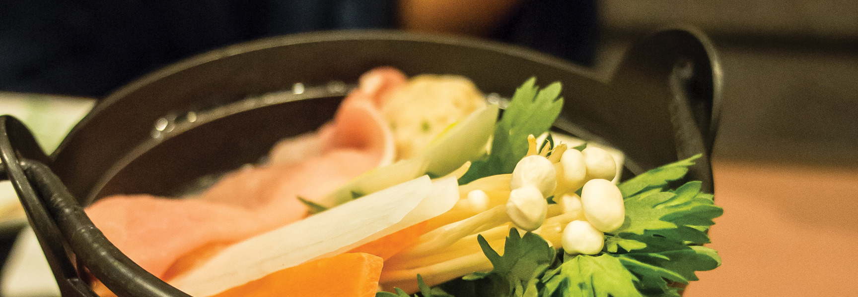 A close-up of a traditional Japanese hot pot, filled with fresh enoki mushrooms, leafy greens, thinly sliced meat, and other vegetables.