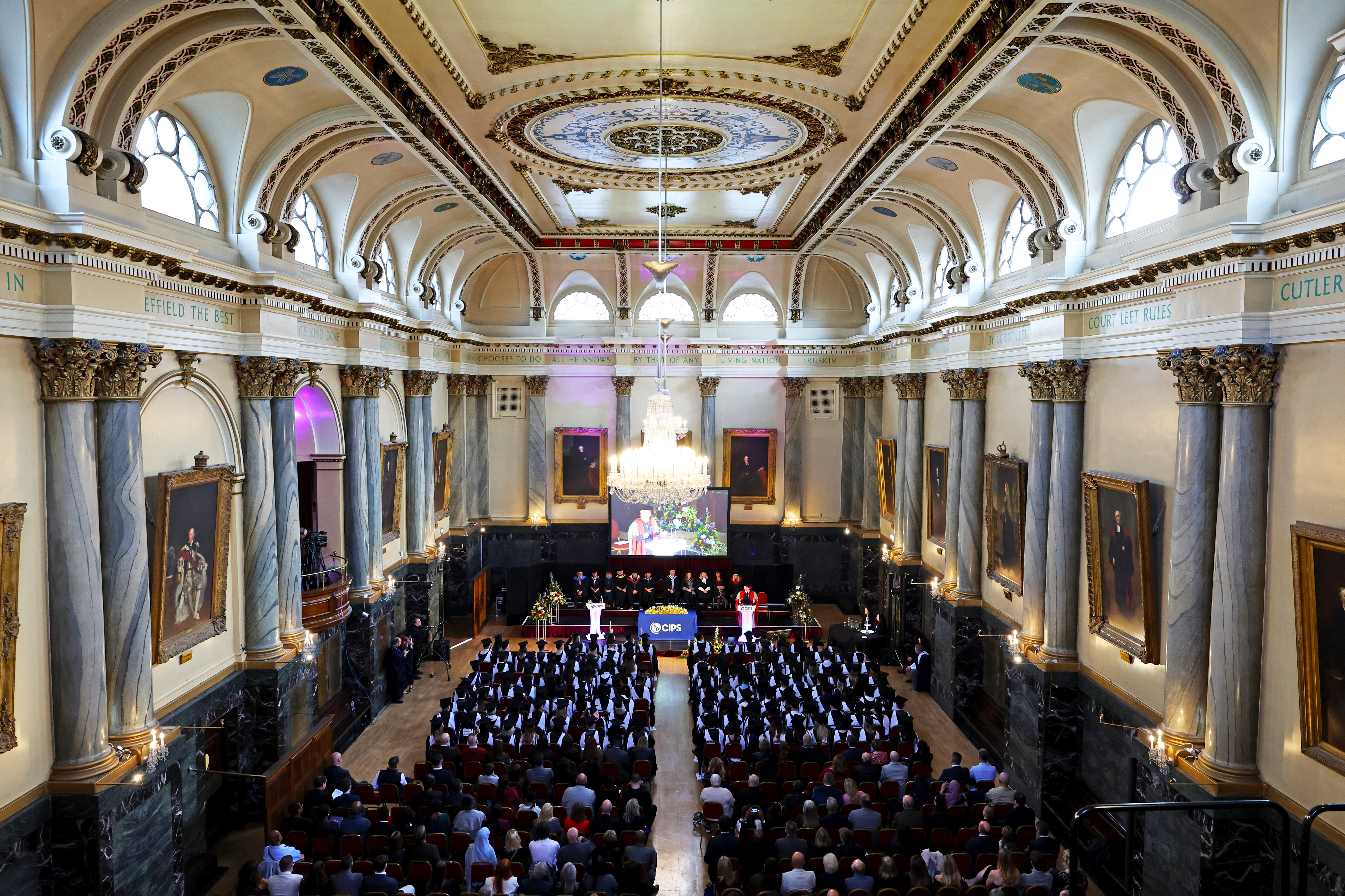 High-angle wide view of a grand graduation ceremony inside a historic hall filled with seated graduates and an audience.