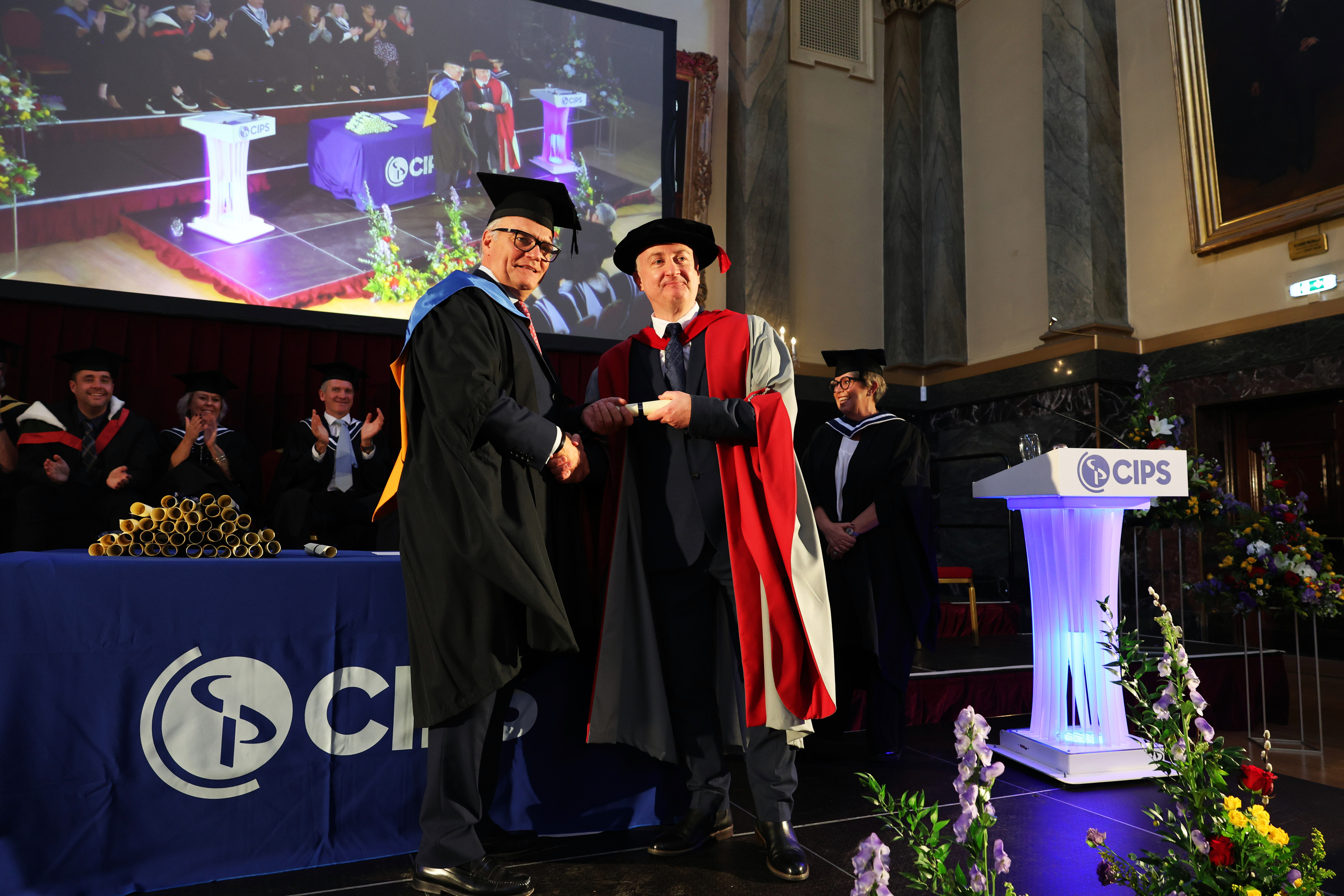 CIPS CEO Ben Farrell shaking hands with a speaker on stage, both dressed in academic robes