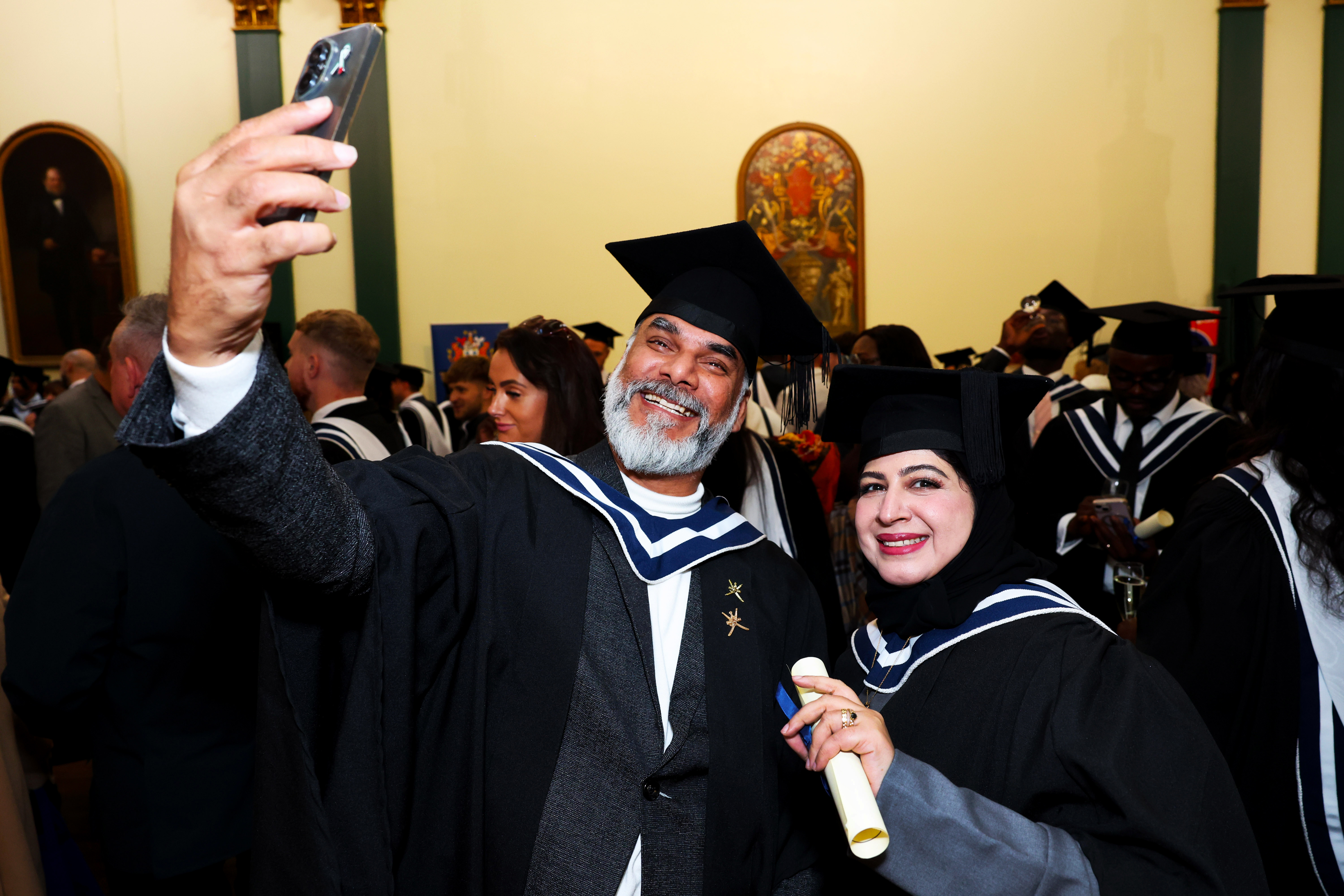 Two graduates in cap and gown pose together for a selfie while proudly showing off their certificate