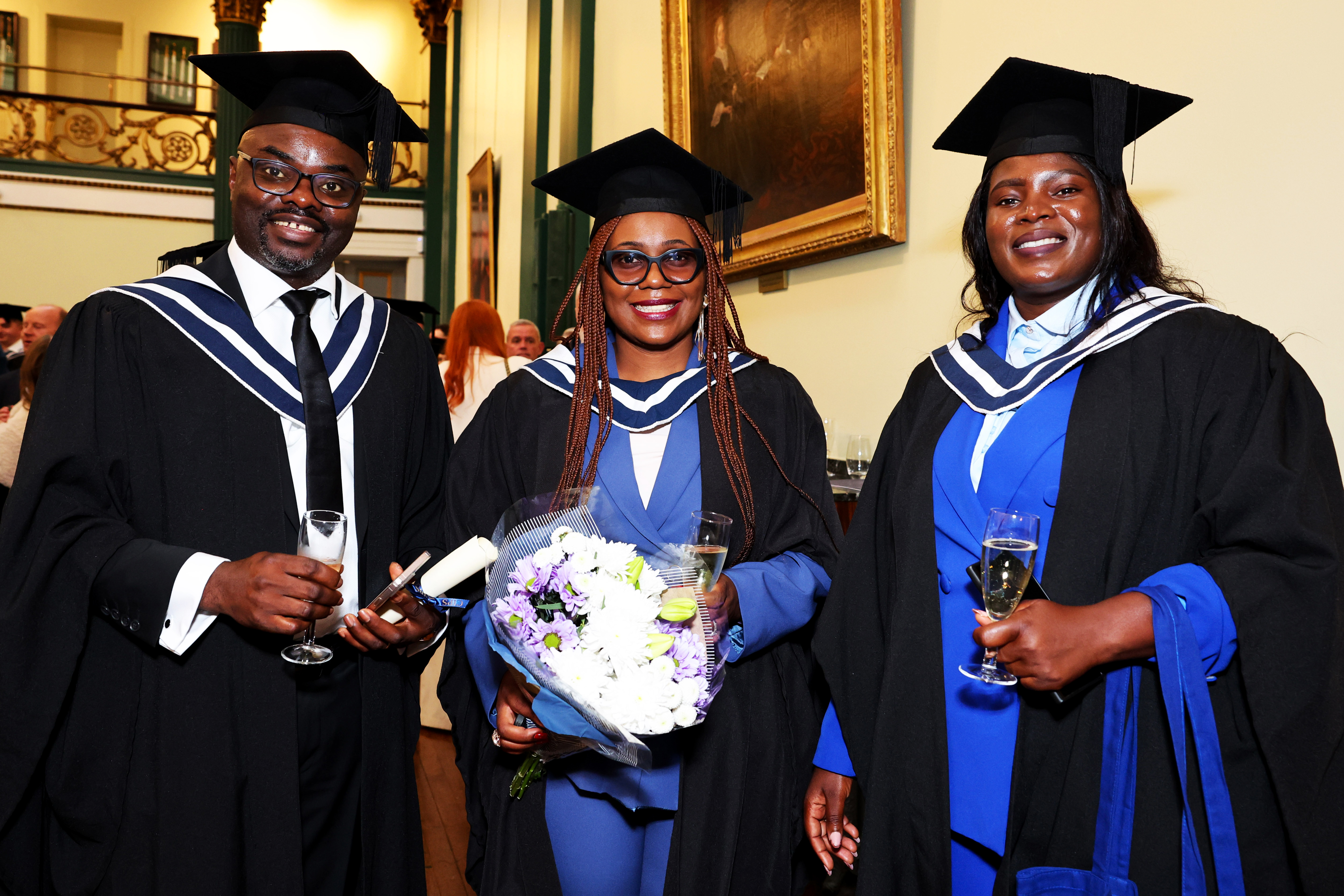 Three graduates in cap and gown (one man, two women) celebrate after the ceremony, holding flowers, drinks and a certificate