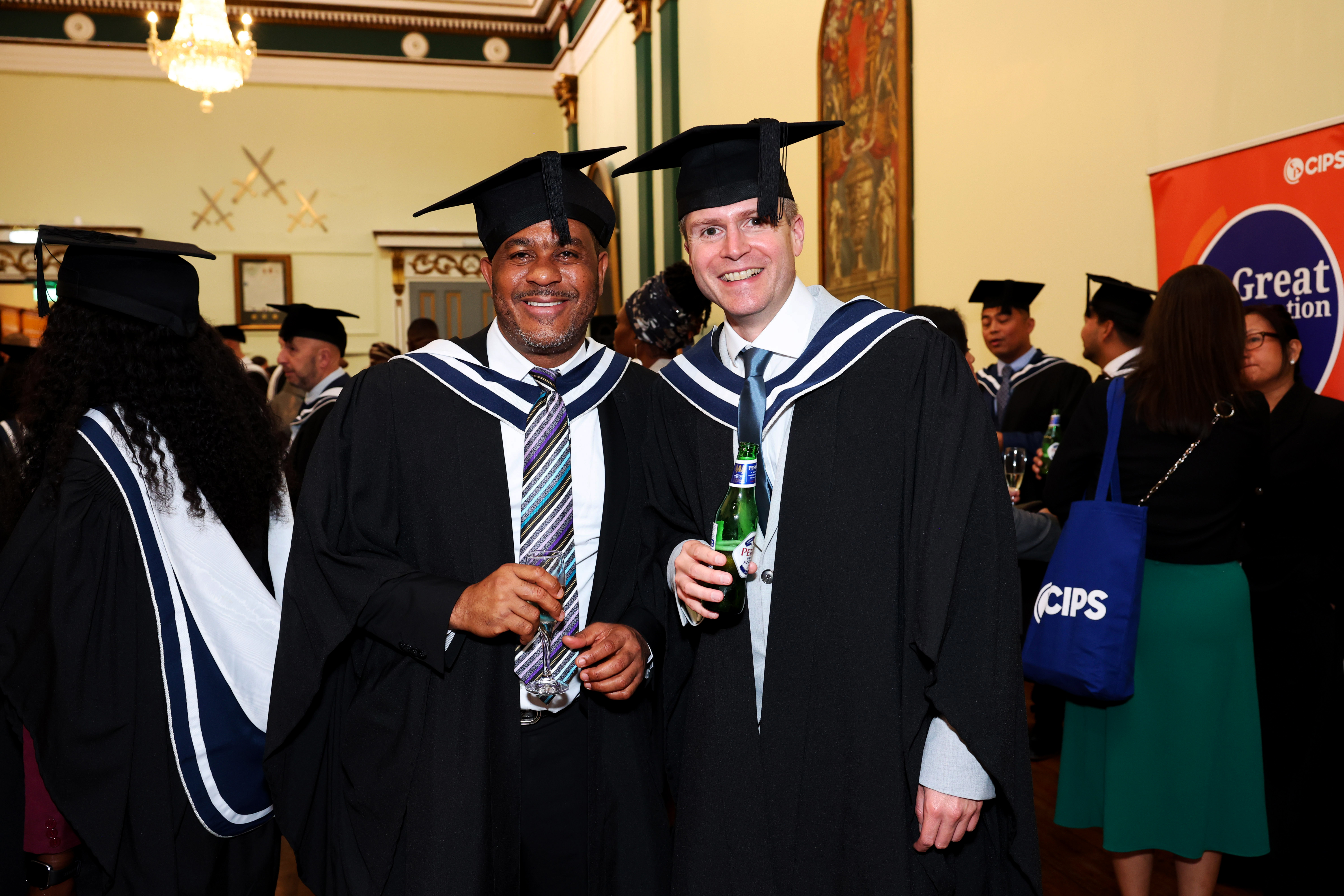 Two men in graduation gap and gown pose side by side holding drinks at the graduation reception