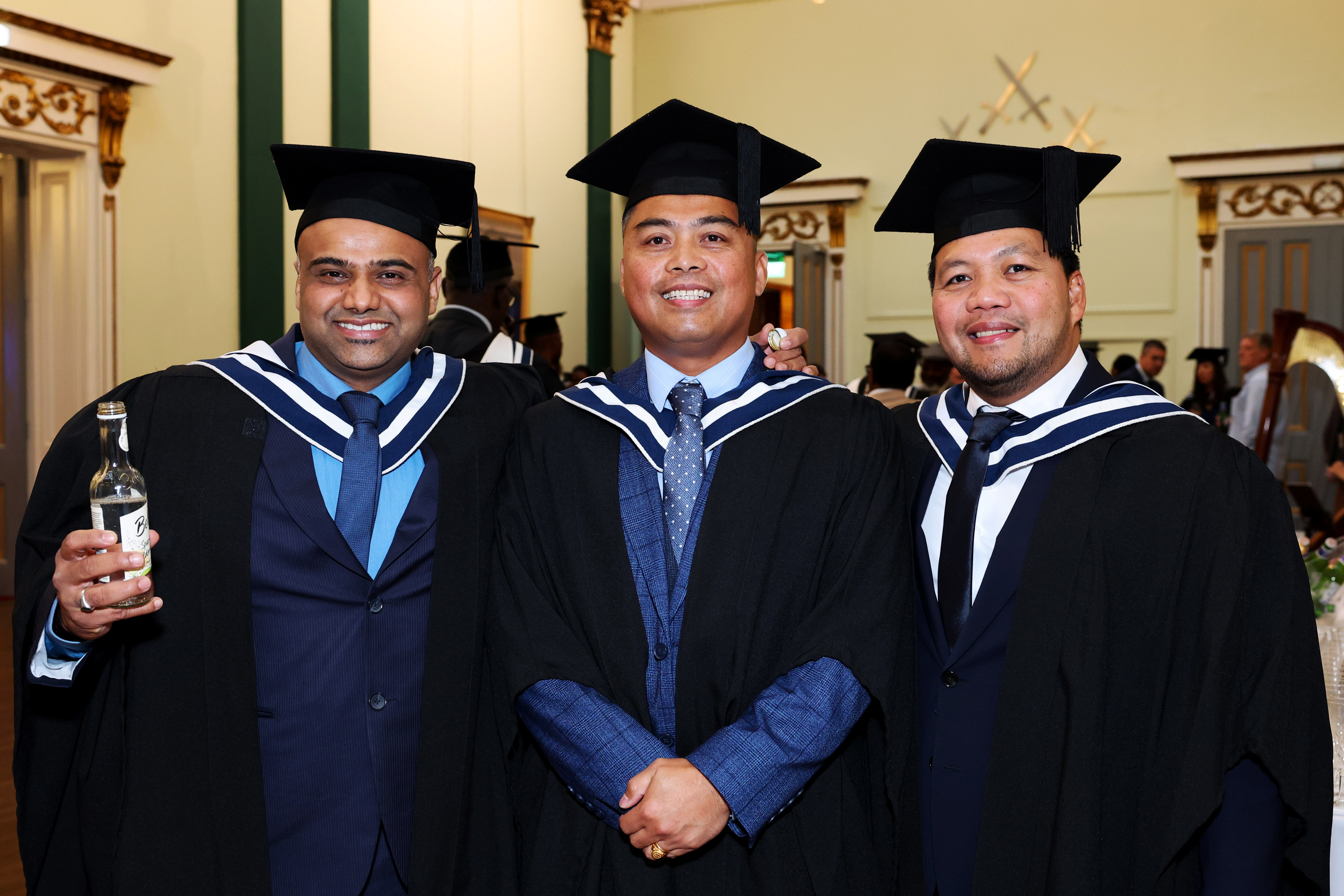 Three male graduates in cap and gown celebrating after the ceremony, holding drinks