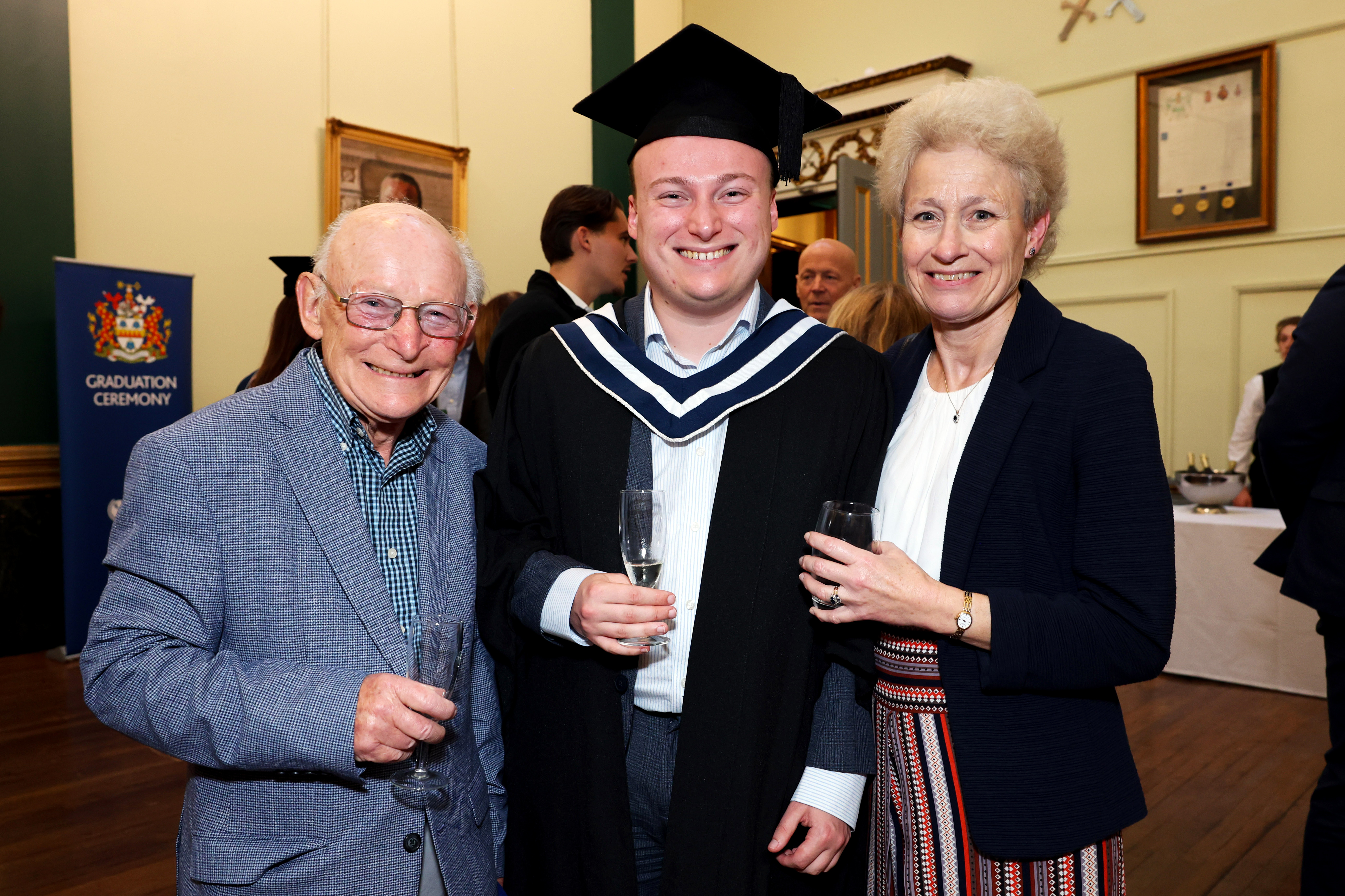 Male graduate in cap and gown posing with two elderly family members, holding drinks