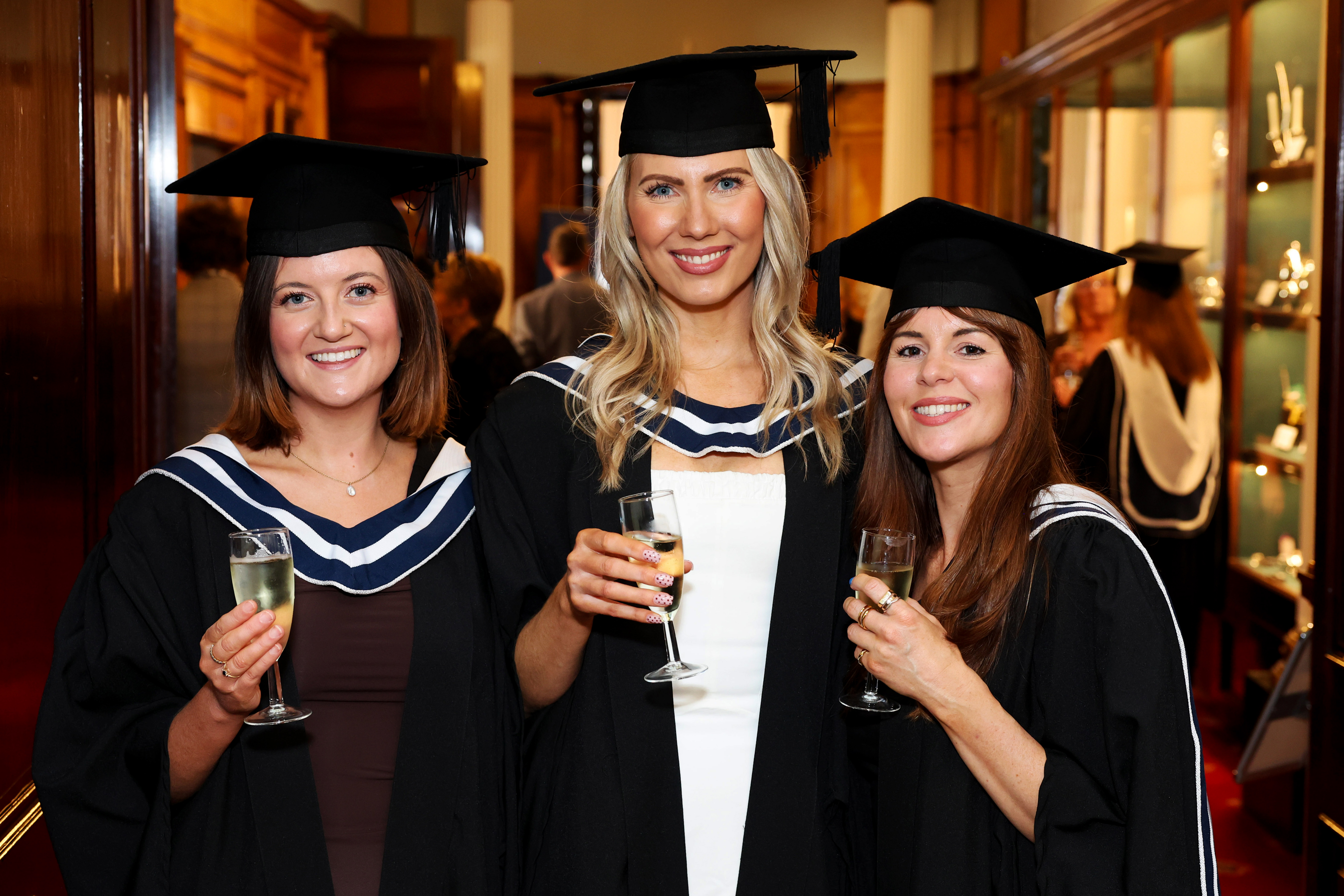 Three female graduates in cap and gown celebrating after the ceremony, holding drinks and showing off their diplomas