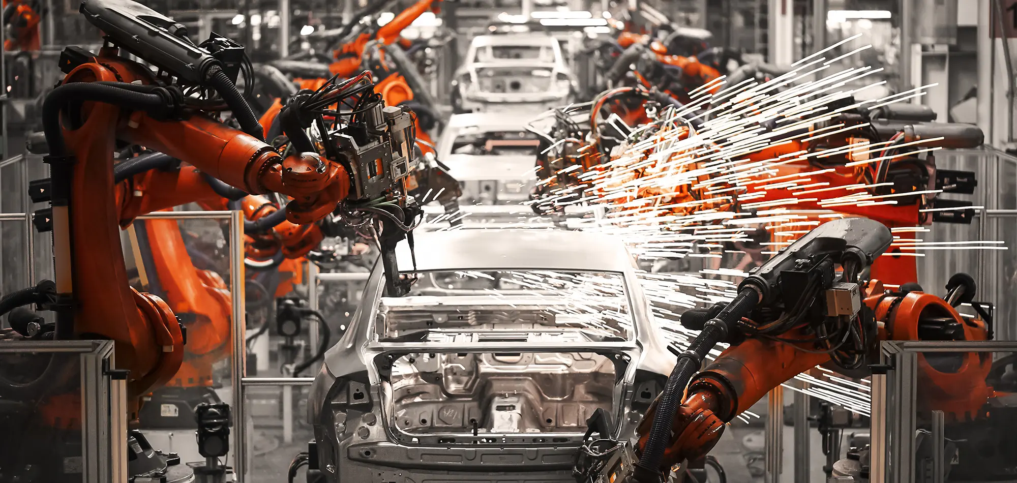 An orange and black robotic arm is actively welding a silver car body on an assembly line, producing a bright shower of sparks. Several other orange and black industrial robots surround the car, working on other parts of the factory floor. The image is predominantly black and white except for the orange color of the robot arms and the bright white sparks.