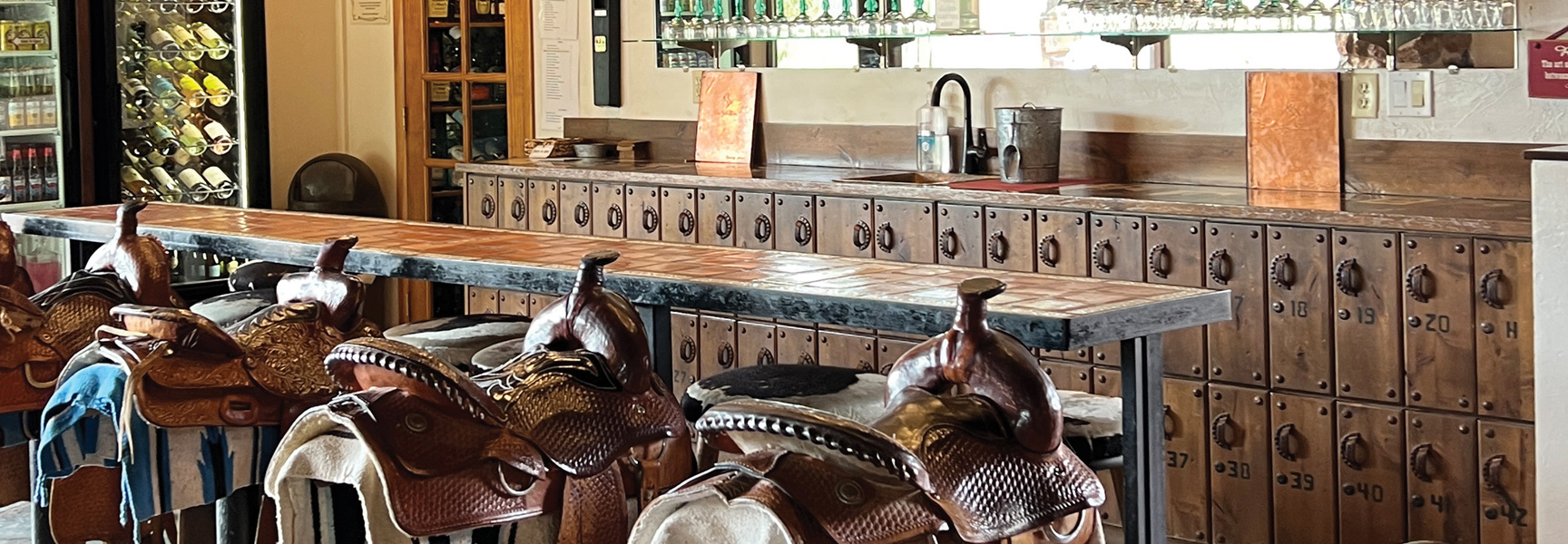 A rustic bar in Arizona with a tiled countertop and leather horse saddles used as bar stools in front of a wall of lockers.