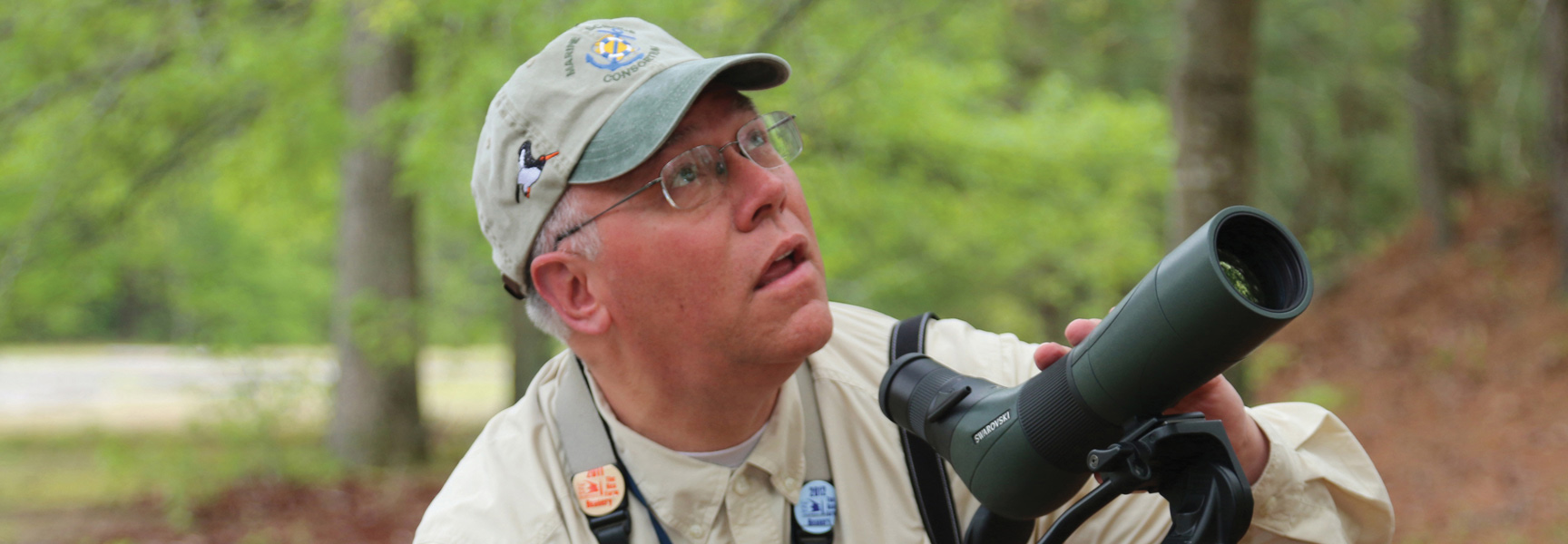 A man in a hat and glasses looks up into the trees while holding a large spotting scope during a birding tour in Virginia.