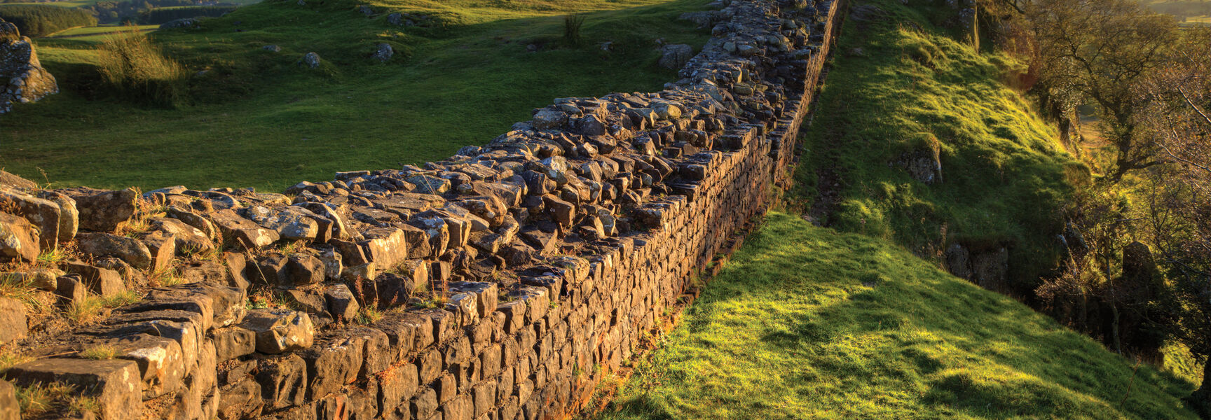 The stone remains of historic Hadrian’s Wall stretch across the rolling green hills of the English countryside under a soft sunset sky.