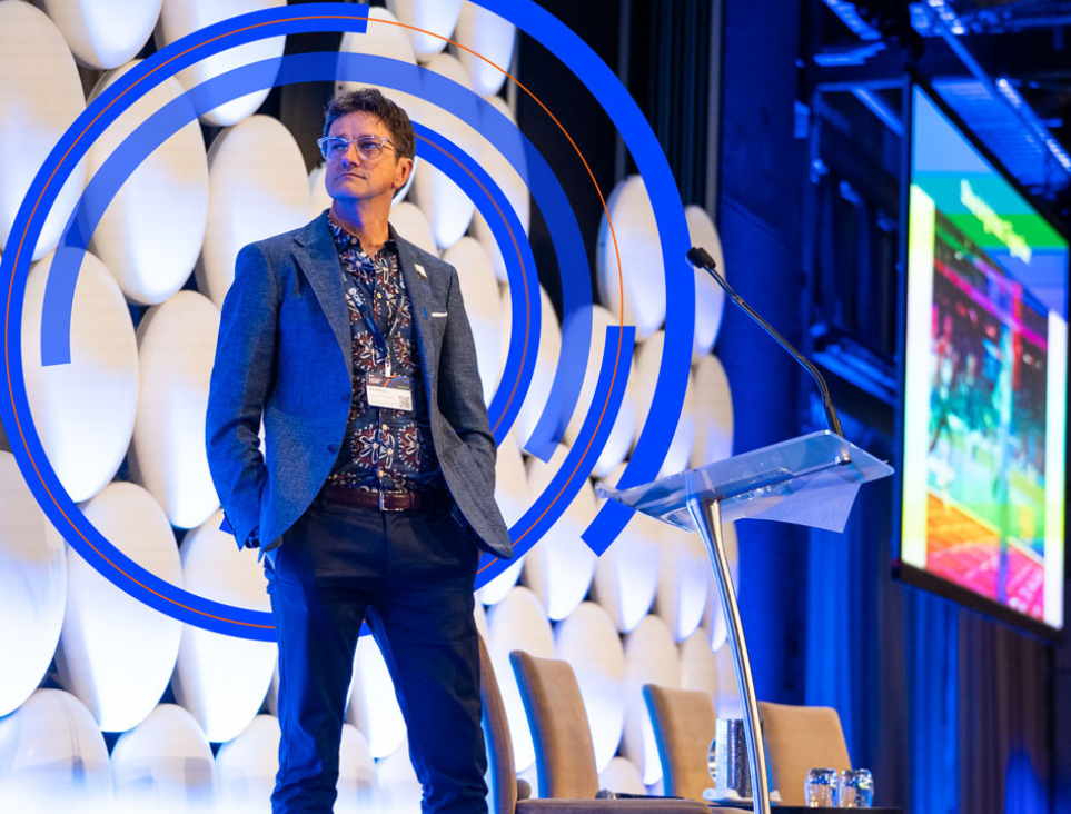 A man in a blue blazer and patterned shirt stands thoughtfully on a conference stage framed by blue circular graphic overlays. A podium and a projection screen are visible in the background against a textured wall of white oval panels.