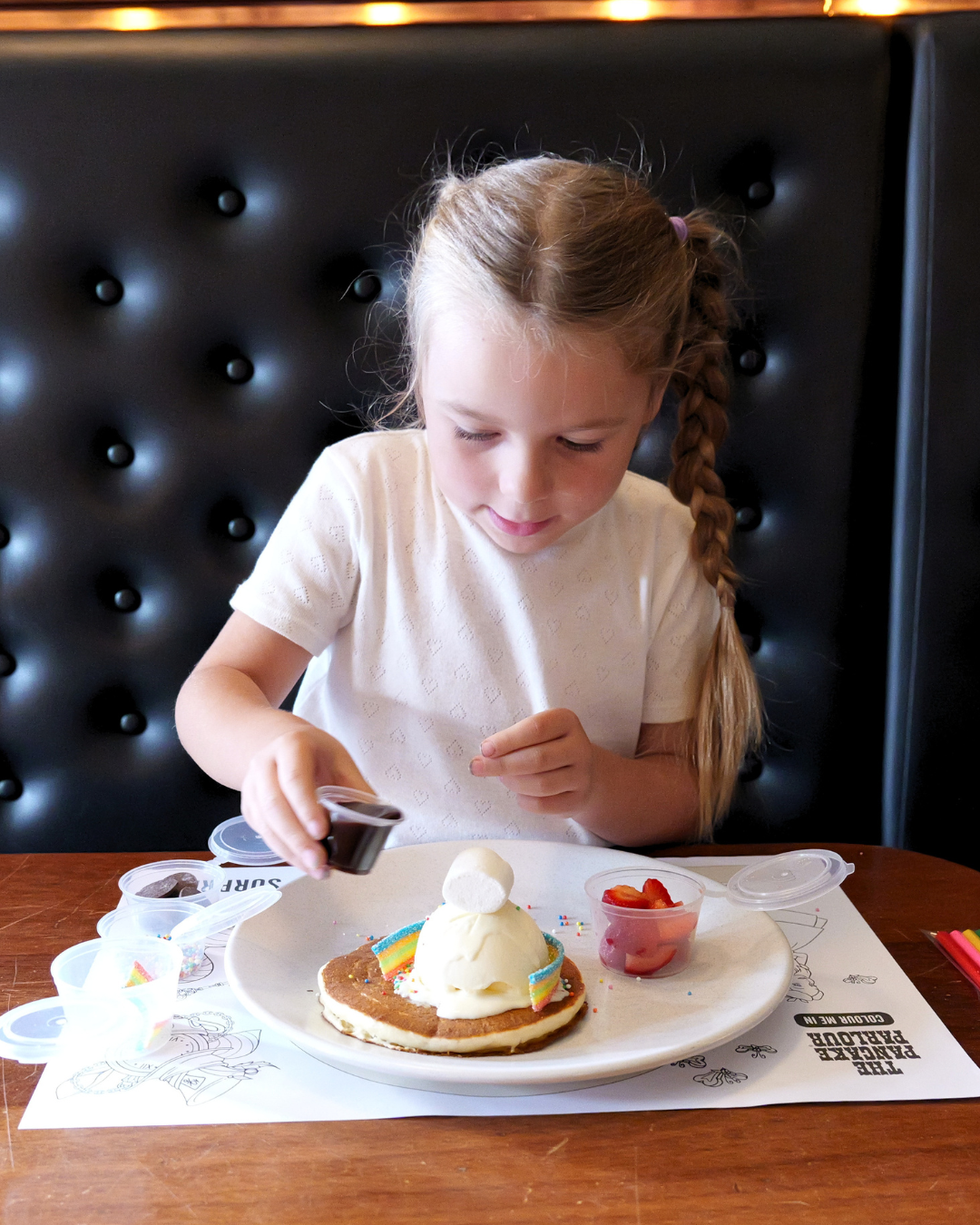 A child sitting in a restaurant booth decorating a pancake topped with ice cream, strawberries, marshmallows and colourful toppings, with small containers of sprinkles and sauce on the table.