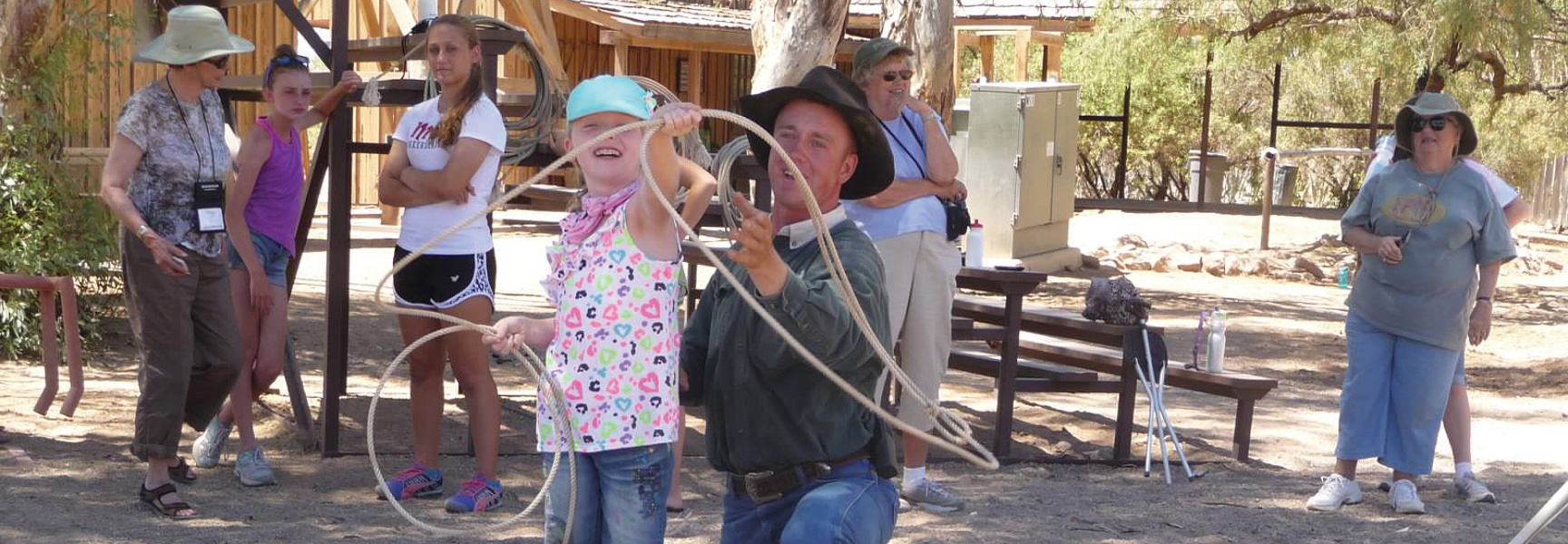 A man in a cowboy hat teaches a young girl how to use a rope lasso while a group of people watches at a ranch in Arizona.
