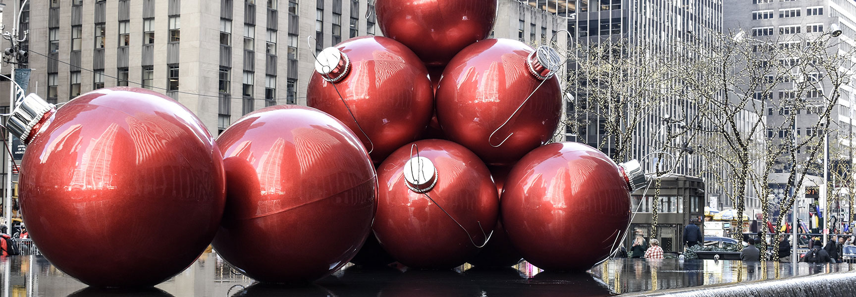 Giant red Christmas ornaments are displayed in a fountain on Sixth Avenue in New York City, with skyscrapers reflecting on their shiny surfaces.