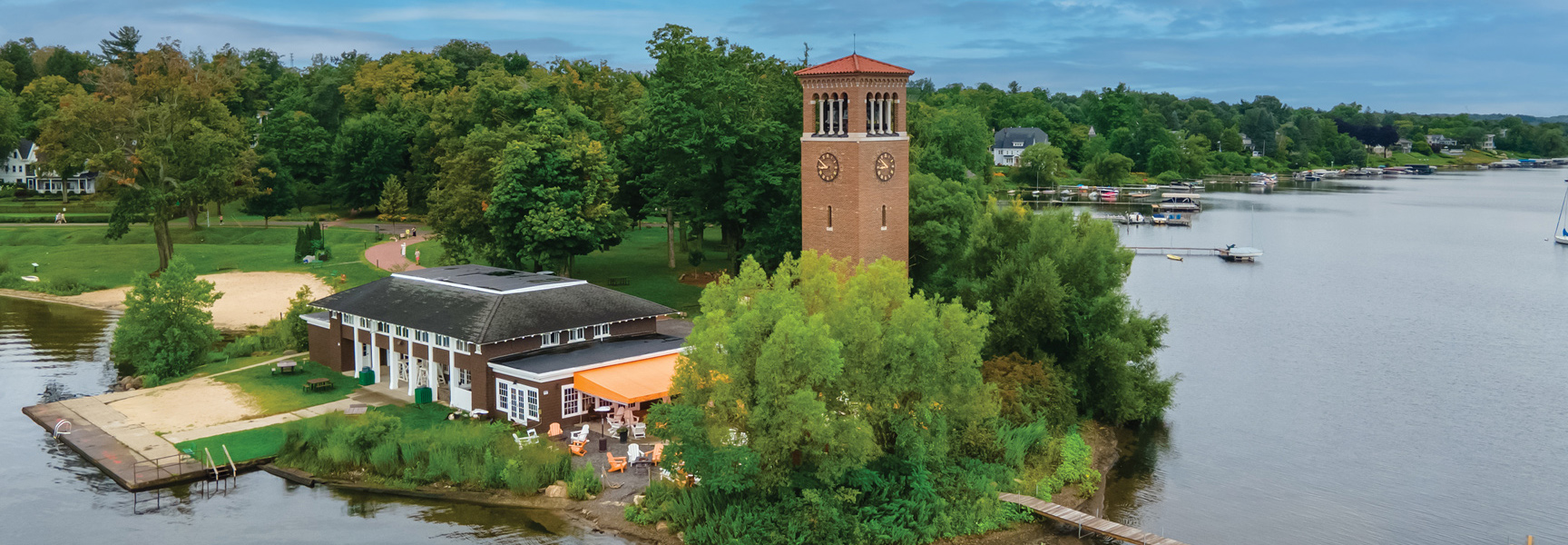An aerial view of the Chautauqua Institution's lakeside village in New York, featuring a prominent brick clock tower surrounded by trees and water.
