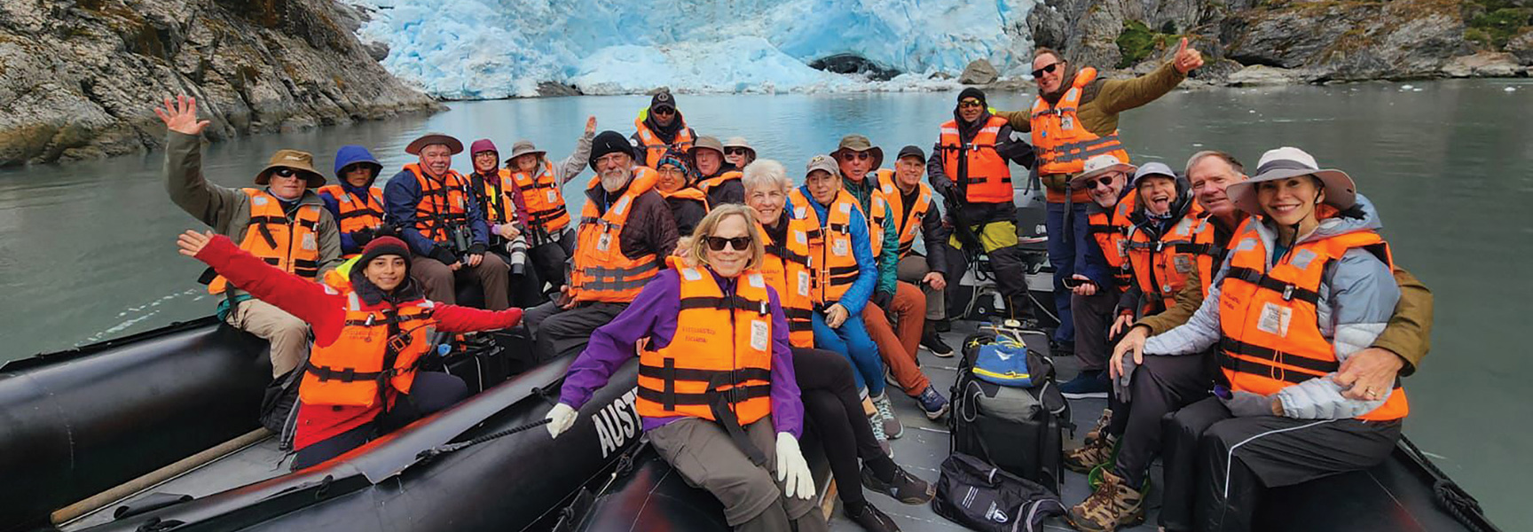 A group of travelers in orange life vests smiles on an inflatable boat in front of a massive glacier in Southern Patagonia, Argentina/Chile.