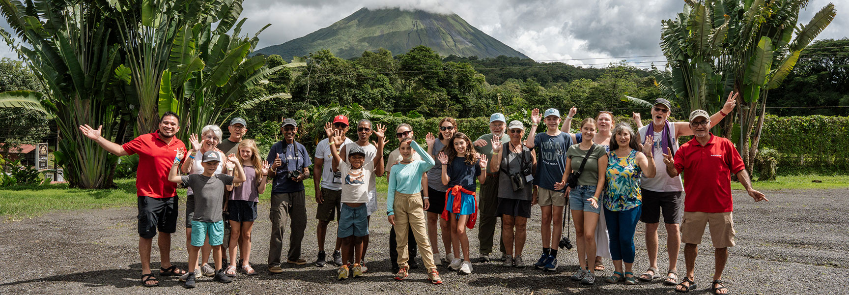 A large group of smiling travelers wave for a photo in front of a volcano and lush tropical plants in La Fortuna, Costa Rica.