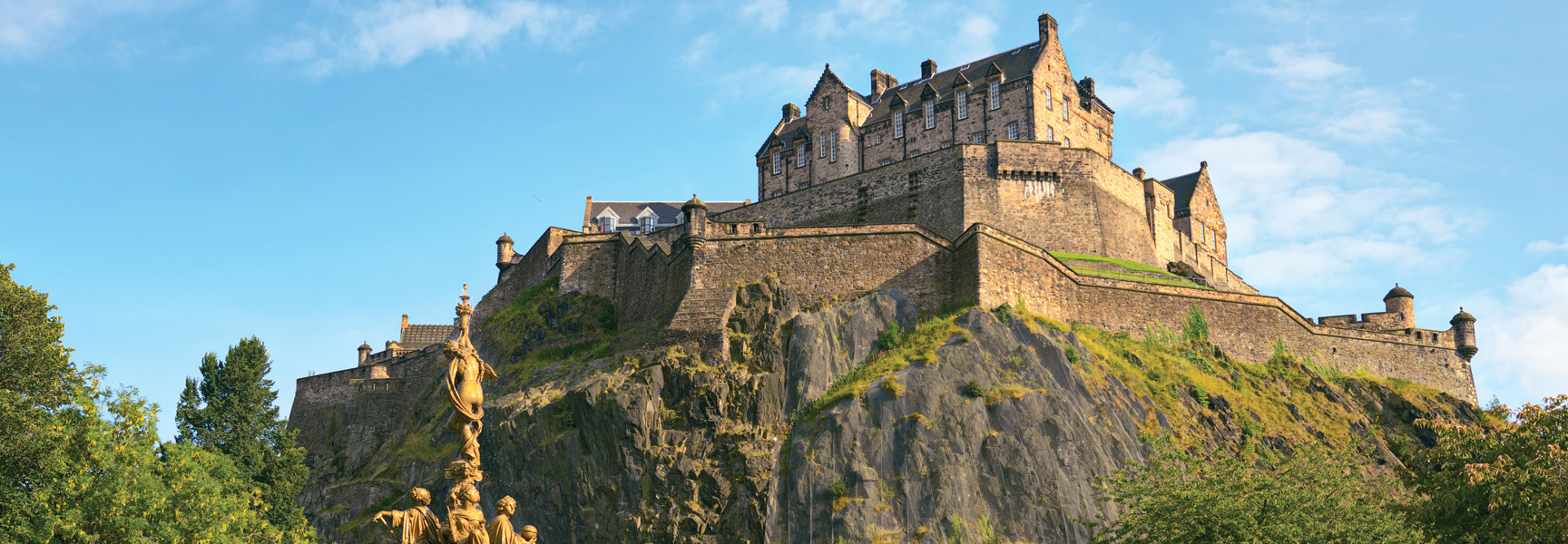 The historic Edinburgh Castle sits atop a large, rocky cliff under a blue sky in Scotland.