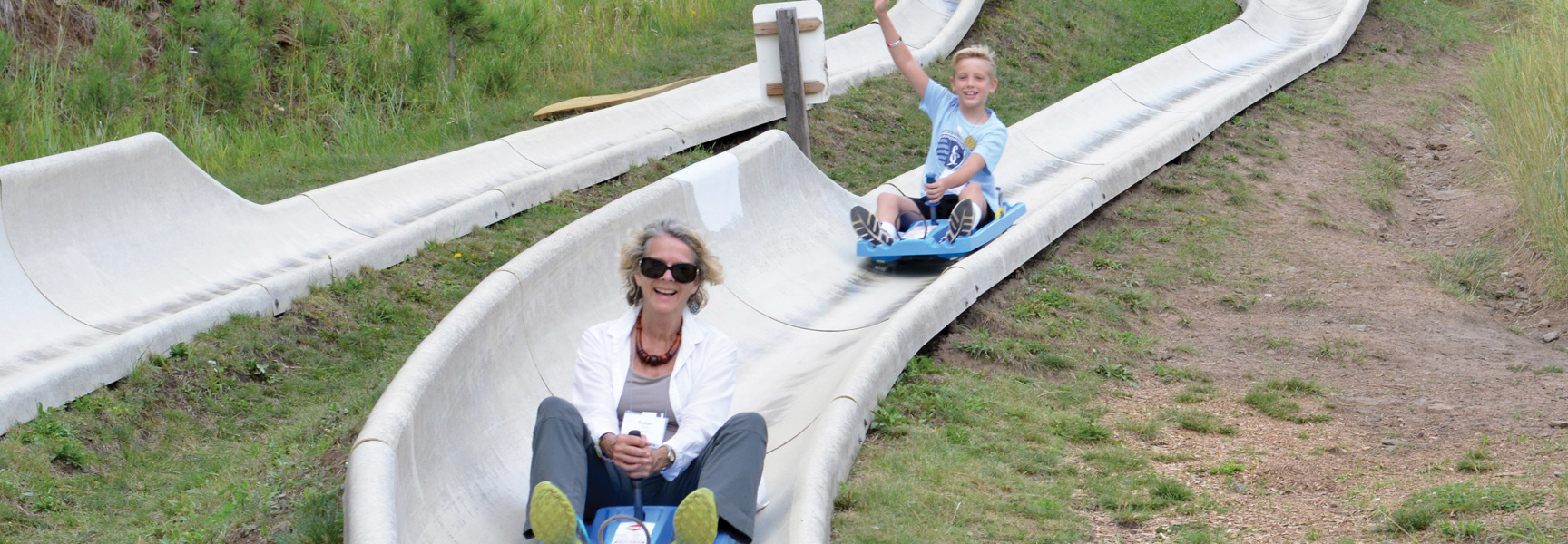 A grandmother and her grandson smile as they race down an alpine slide on a grassy mountainside in Montana.