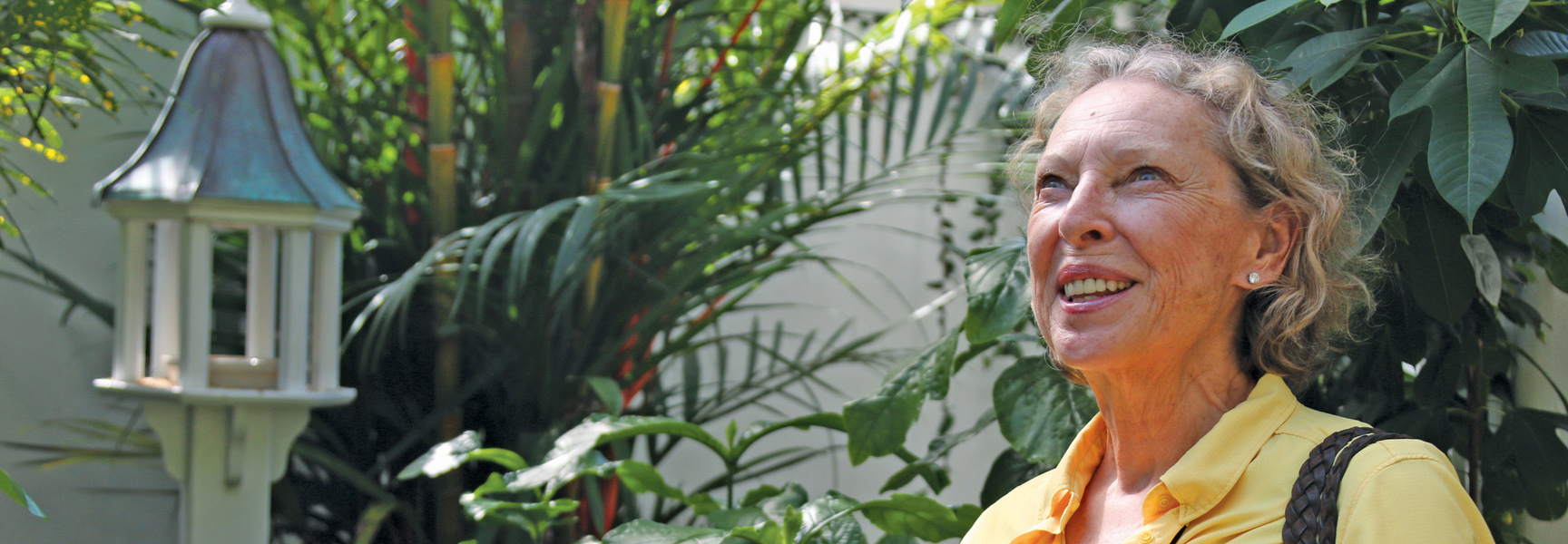 A smiling woman in a yellow shirt stands in a lush Key West, Florida garden next to a birdhouse.