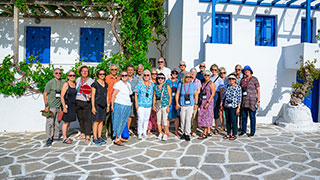 A group of travelers poses on a stone patio in front of a whitewashed building with blue shutters and windows in Paros, Greece.