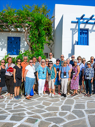 A group of travelers poses on a stone patio in front of a whitewashed building with blue shutters in Paros, Greece.