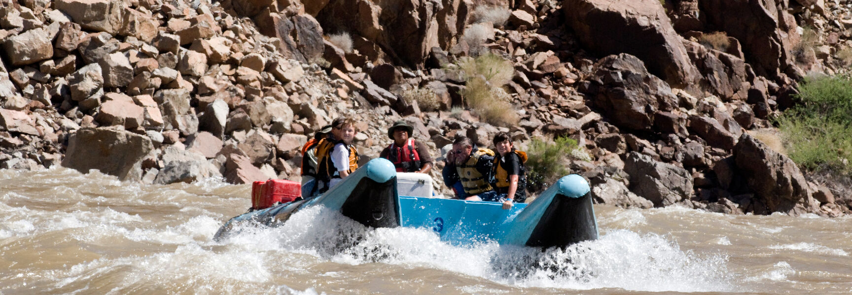 A family navigates river rapids in a blue raft along the rocky canyon walls of Zion National Park.