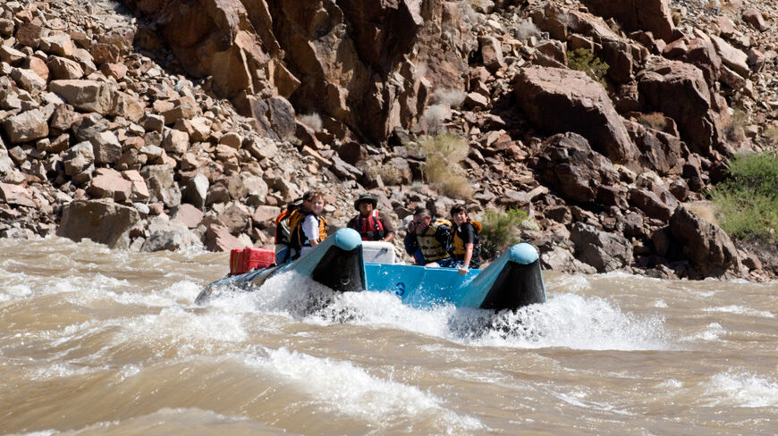 A family navigates river rapids in a blue raft along the rocky canyon walls of Zion National Park.