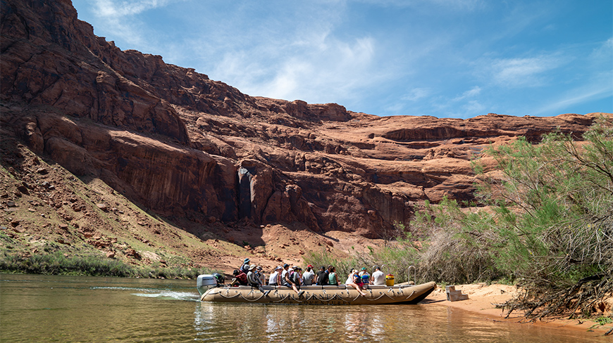 A motorized raft full of people navigates a river at the bottom of a massive red rock canyon in Arizona on a sunny day.