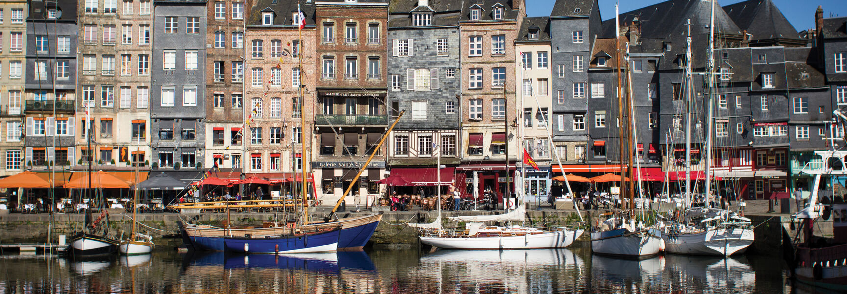 Sailboats dock in a historic harbor in Normandy, France, lined with tall narrow architecture and waterfront cafes reflected in the calm water.