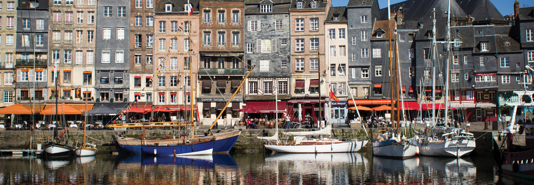 Sailboats are docked in a harbor in front of a row of colorful, narrow buildings in a port in Northern France.