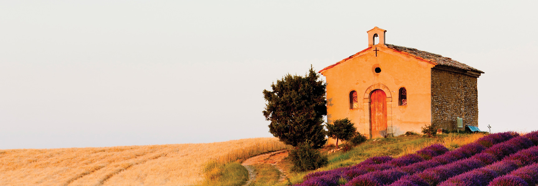 A small stone chapel in Provence, France sits on a hill between a field of golden wheat and rows of purple lavender.