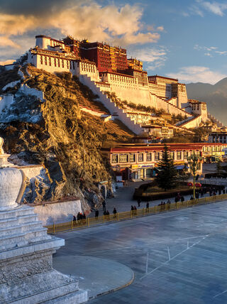 The magnificent Potala Palace in Tibet rises above a white stupa and city square against a backdrop of Himalayan mountains at sunset.