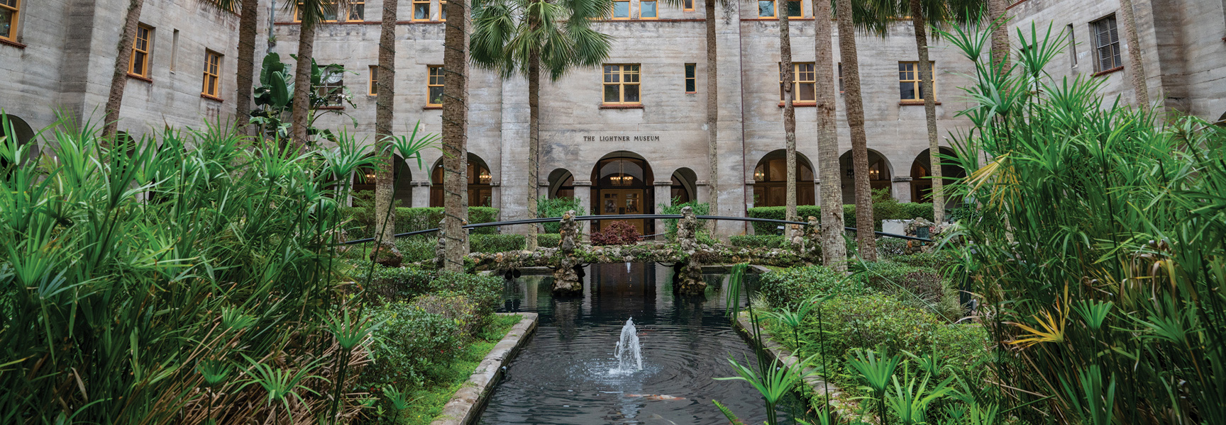 The lush courtyard of the Lightner Museum in Florida features a central pond with a small fountain, a stone bridge, and many palm trees.