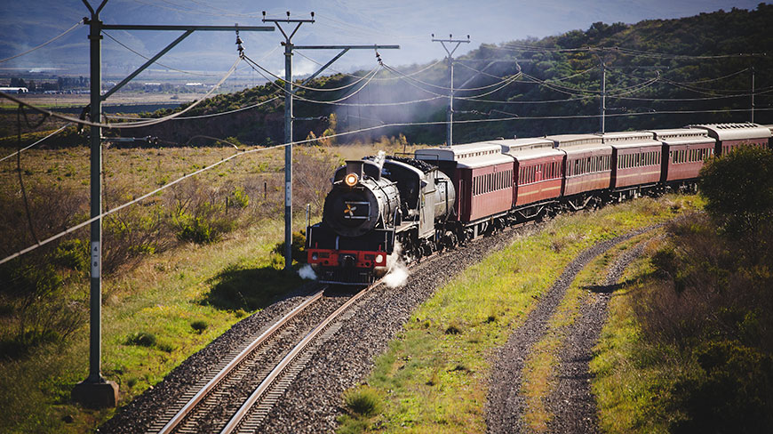 A Rovos Rail steam train journeys through the scenic Southern Africa countryside along a winding track.