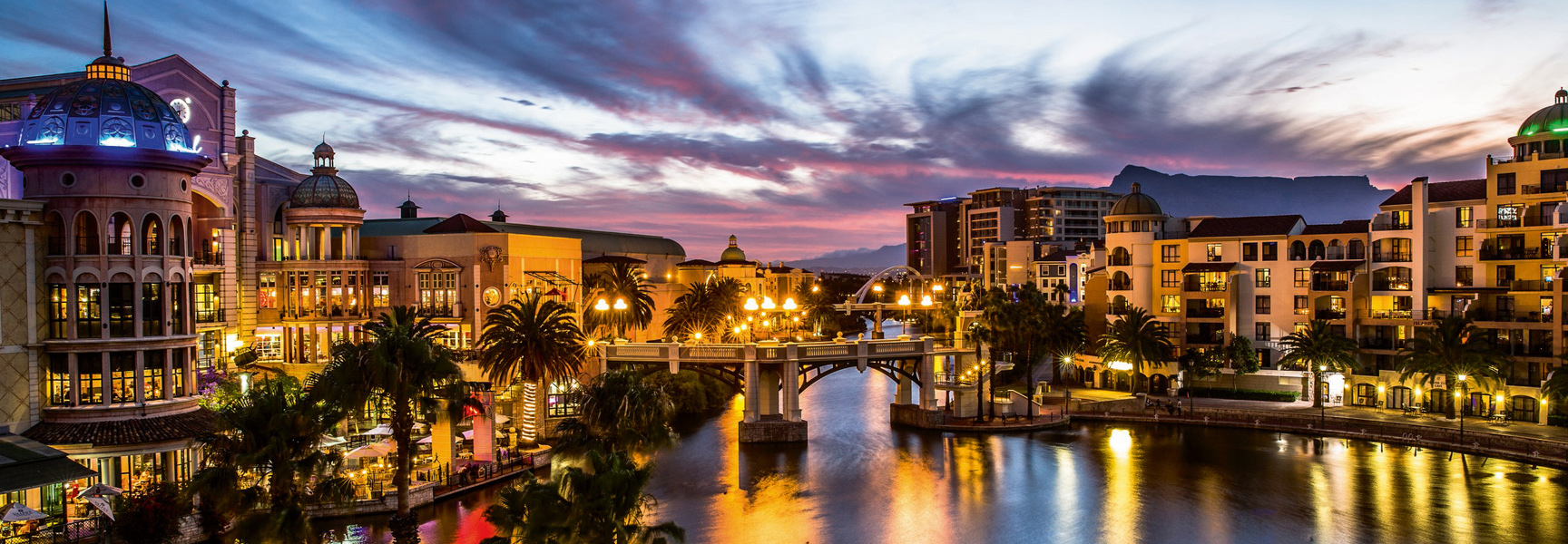 Ornate, illuminated buildings line a canal at the Cape Town waterfront in South Africa at sunset, with Table Mountain in the distance.